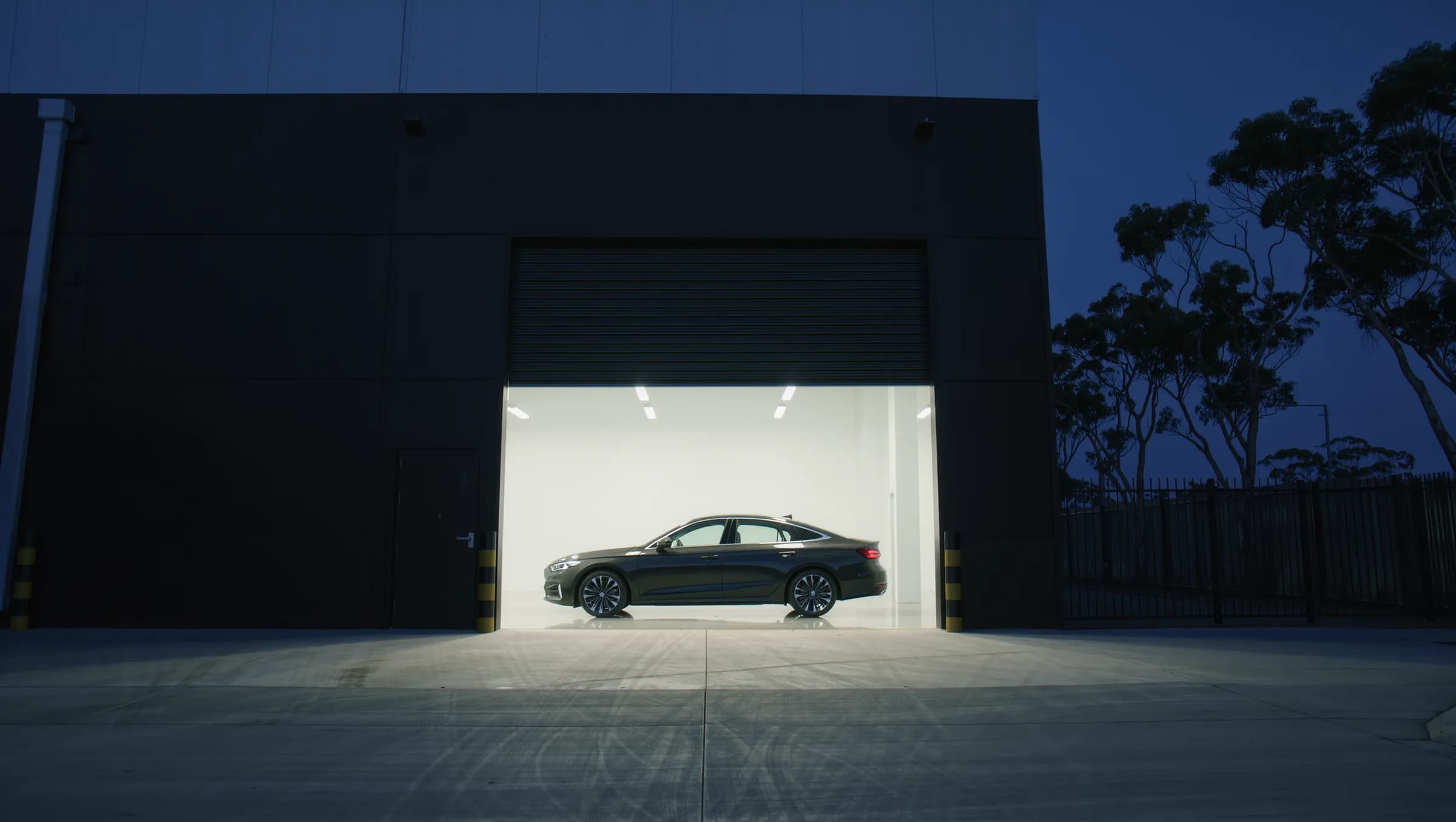 A dark workshop building at blue hour with its roller door open, a sedan lit inside and gum trees beside it.
